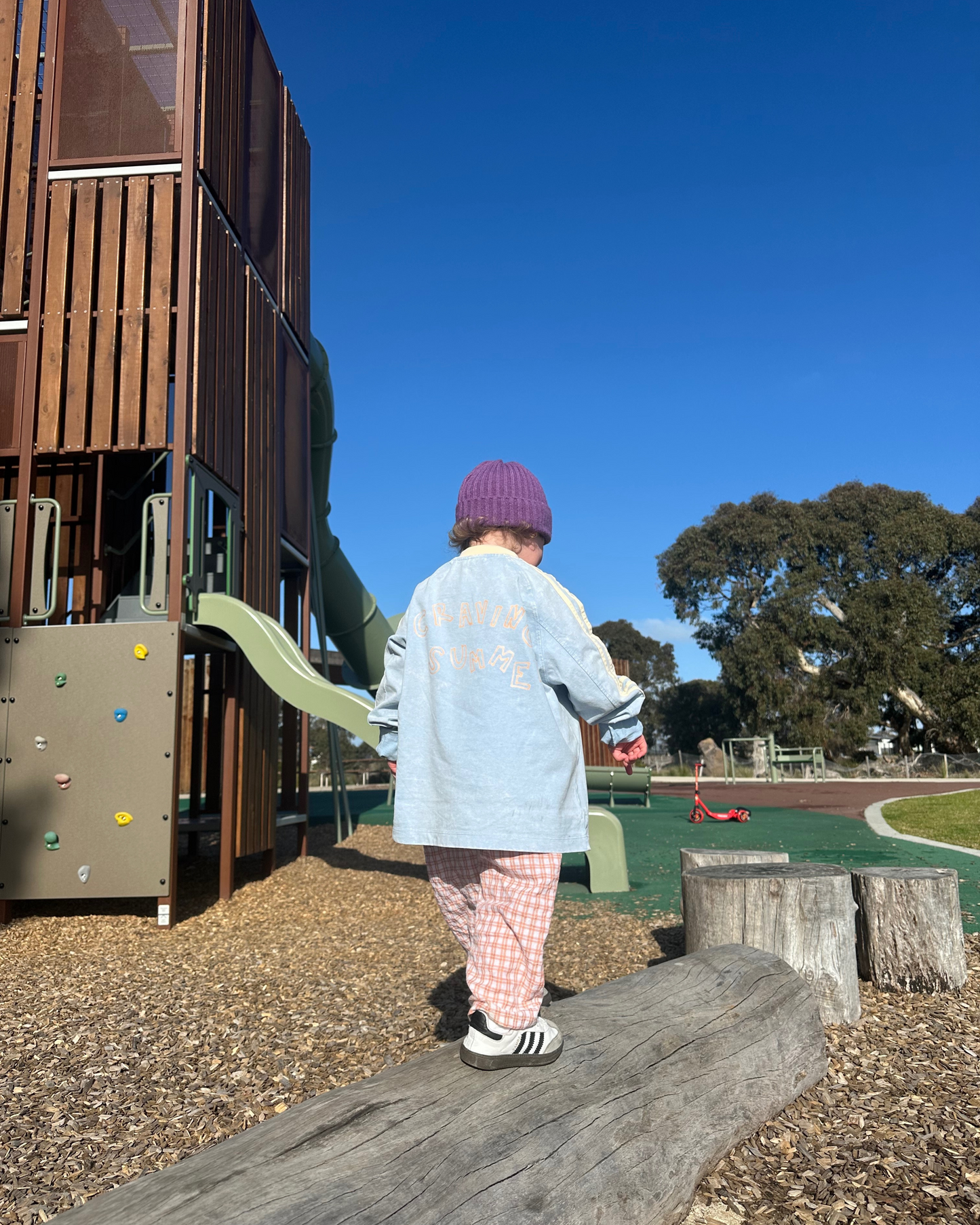 Child standing on a log at a playground with a clear blue sky wearing soltai the labels new collection