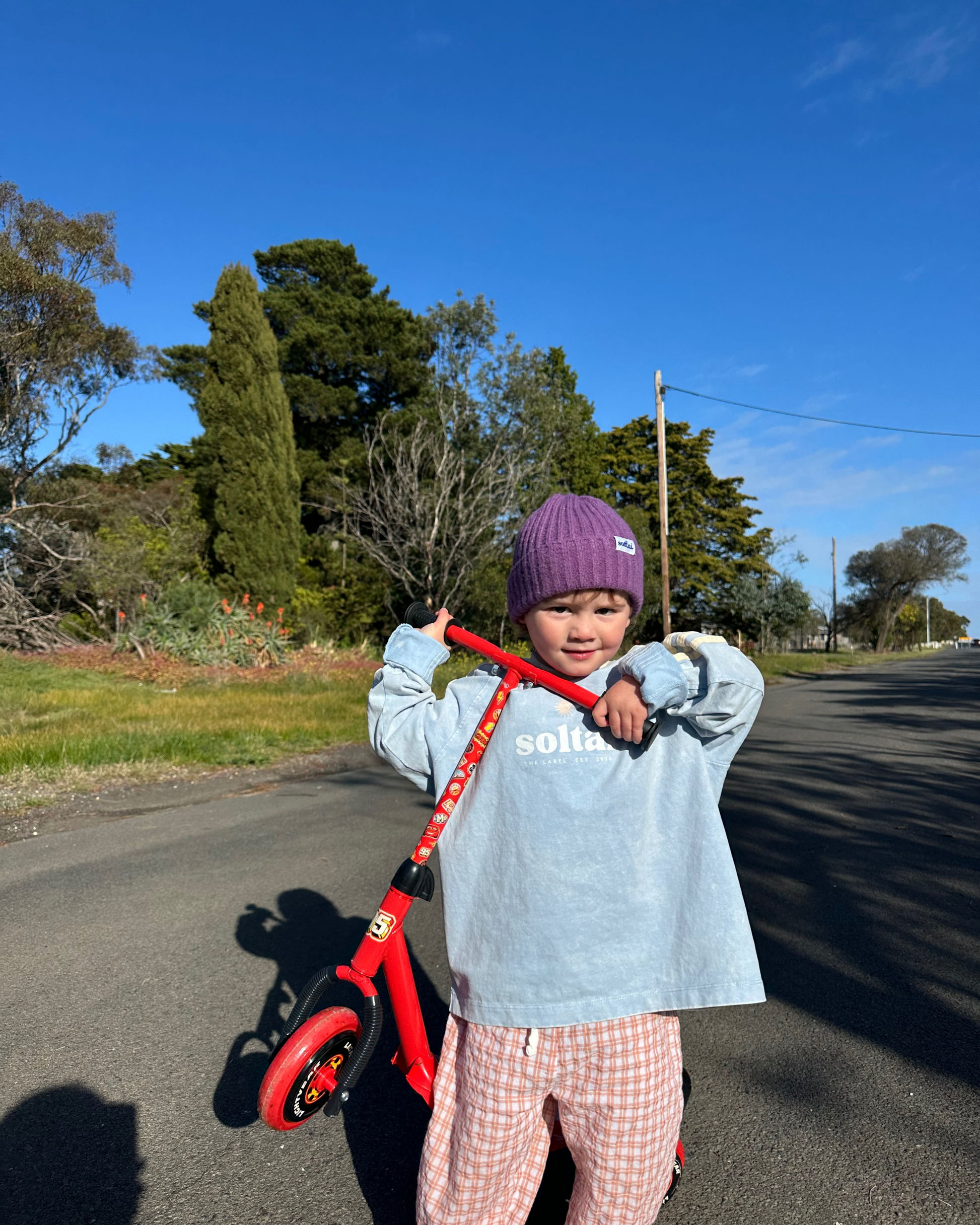 Child holding a red scooter on a road with trees and blue sky in the background wearing soltai the label spring outfits