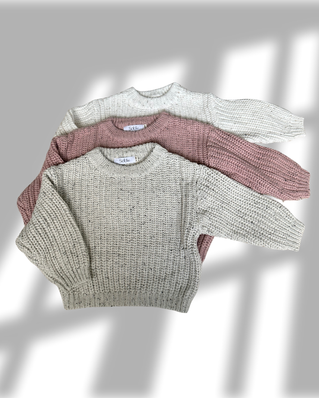Three knit sweaters in white, pink, and gray on a light background