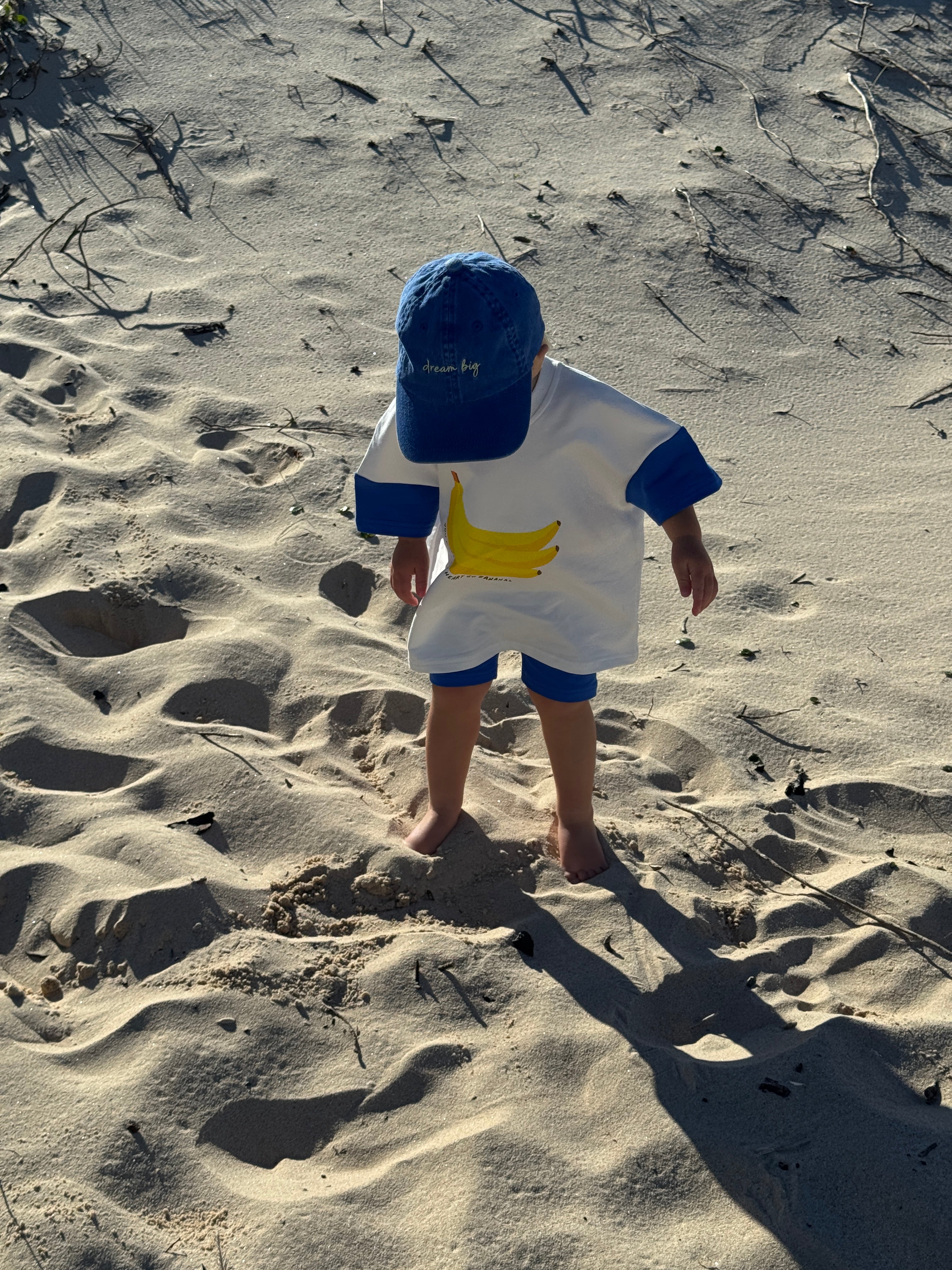 toddler standing on a sandy beach with footprints around, wearing a bright blue playful outfit with a banana print on the front and cobalt blue bike shorts on s summer day in australia