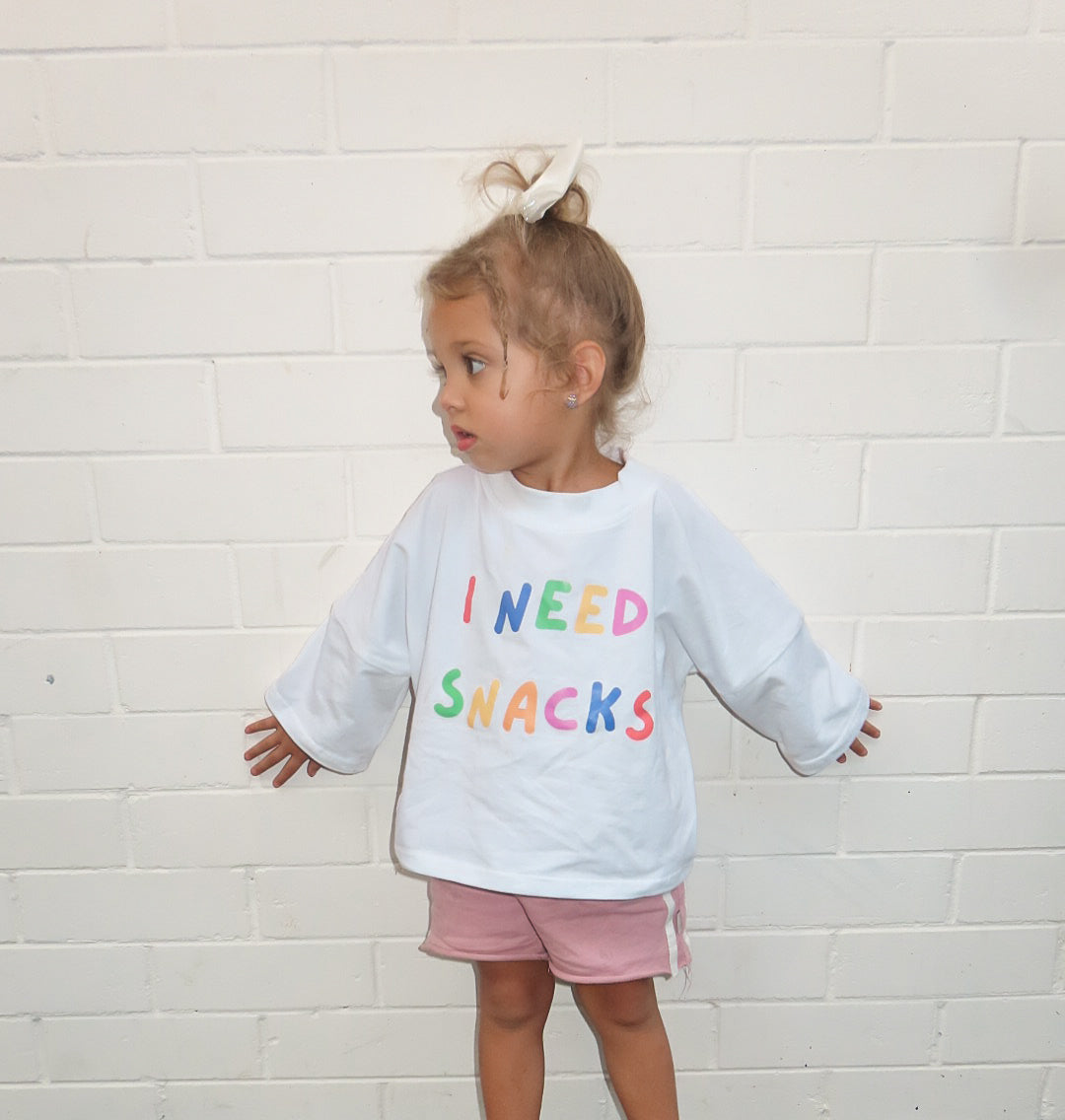 Child wearing a white tee shirt with colourful  i need snacks text standing against a white brick wall. perfect for  a unisex outfits on a nice day in melbourne