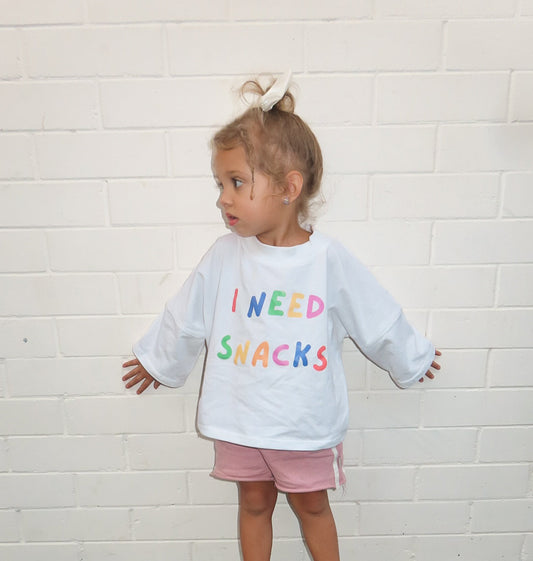 Child wearing a white tee shirt with colourful  i need snacks text standing against a white brick wall. perfect for  a unisex outfits on a nice day in melbourne