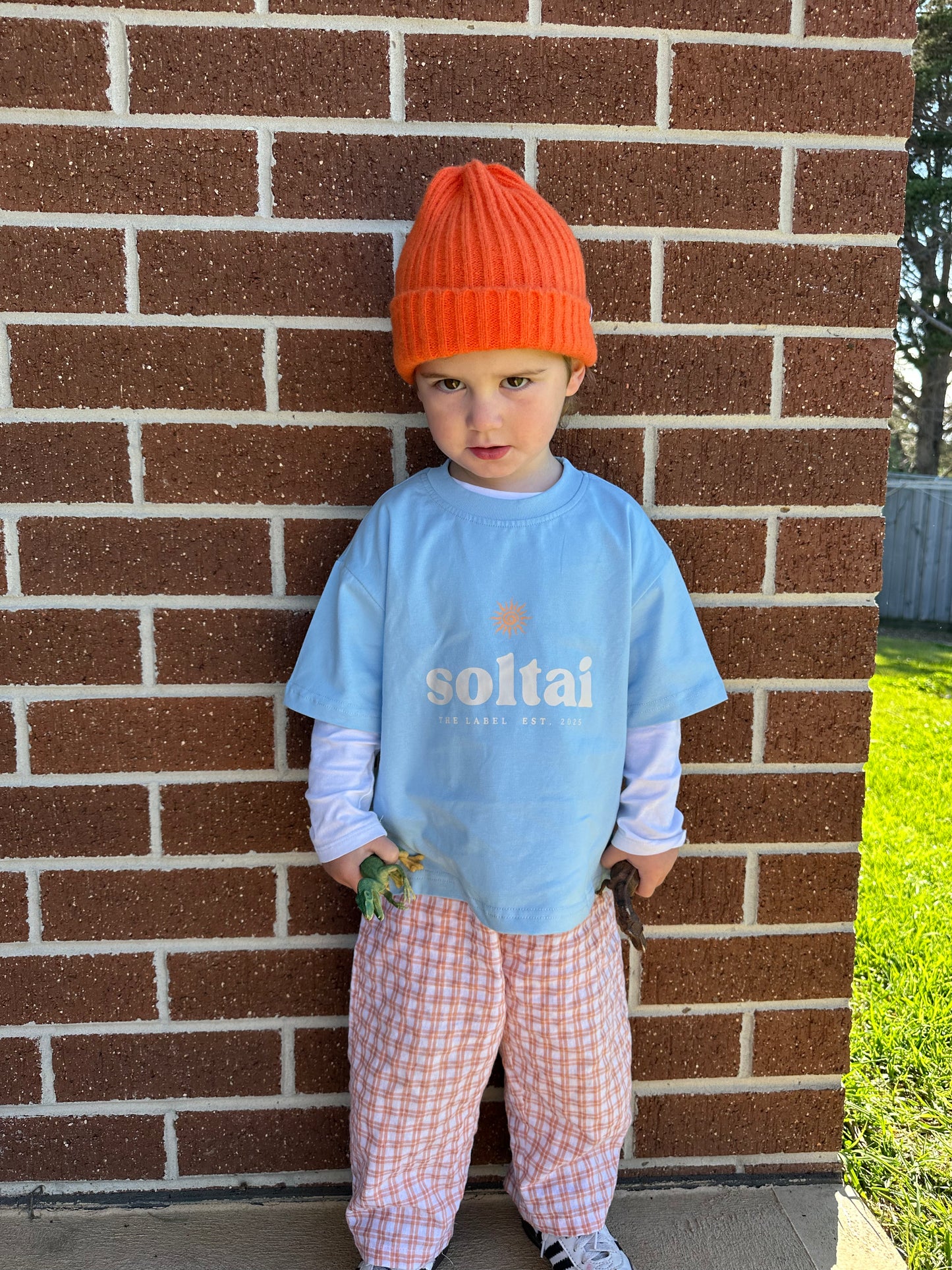 Child wearing a blue shirt with 'soltai' text, orange beanie, and plaid pants standing against a brick wall.