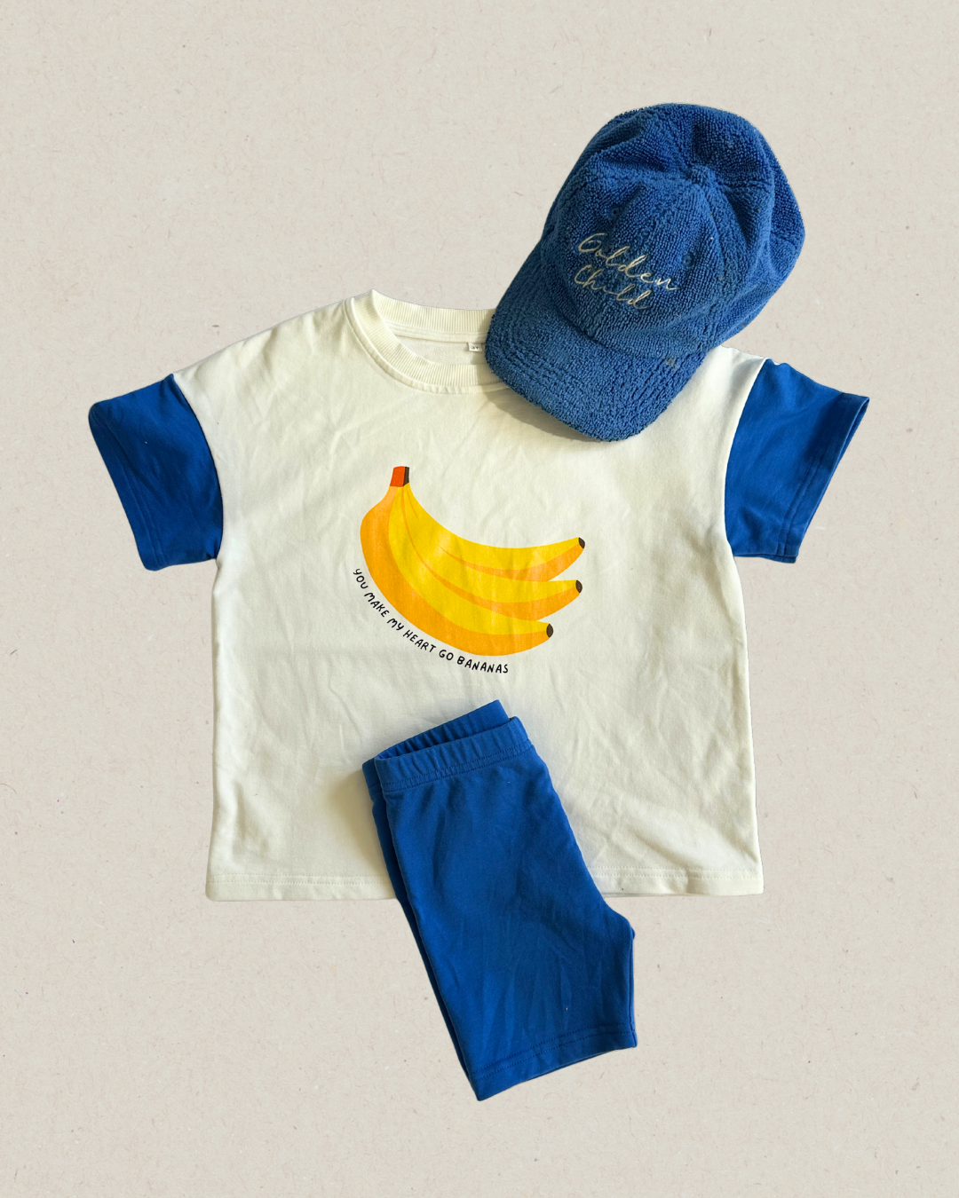 A set of children's clothing consisting of a white t-shirt with a banana print and blue shorts, paired with a blue hat, bright fun and playful kids outfits