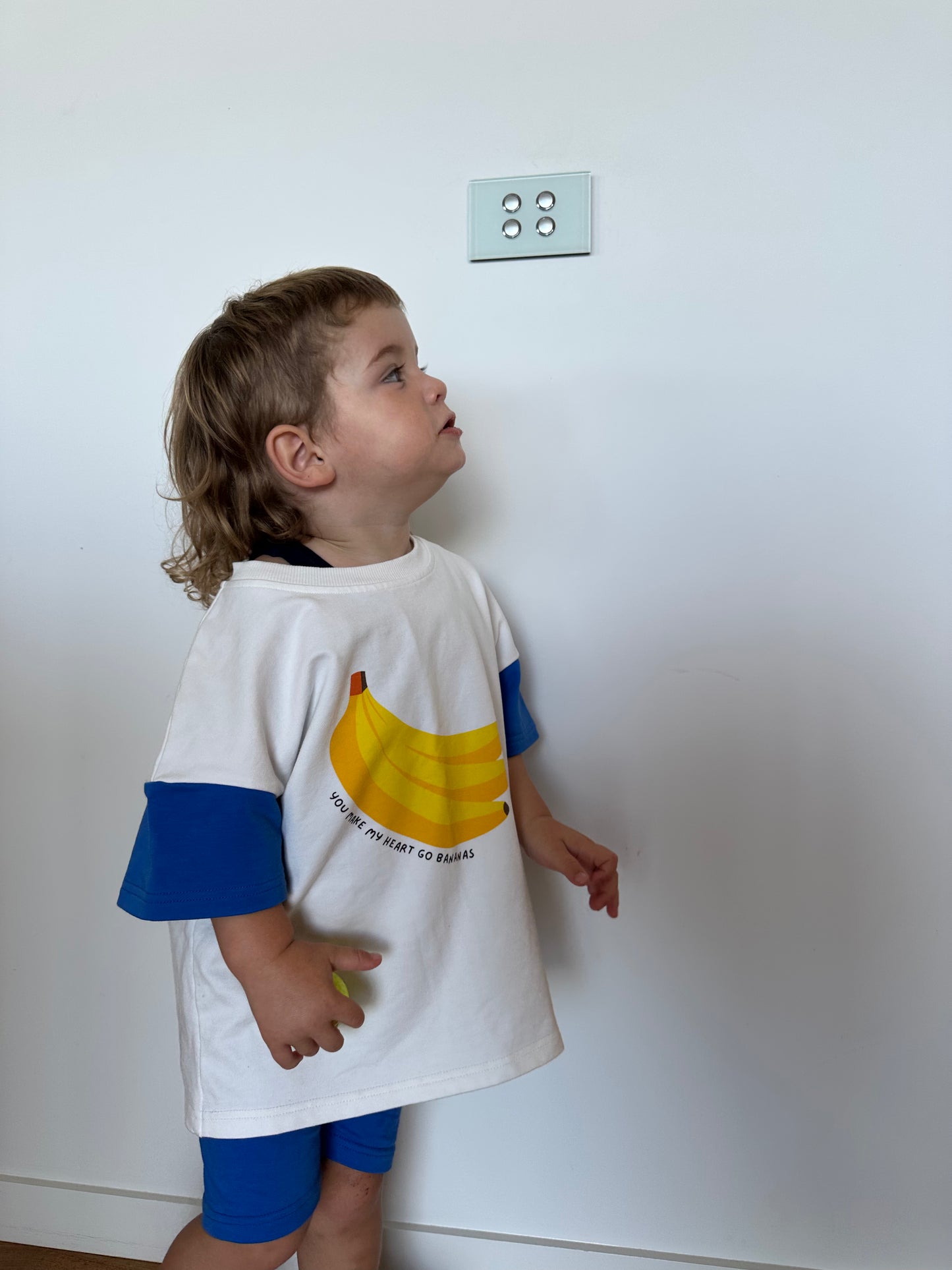 Child wearing a white t-shirt with a banana graphic and text, standing against a plain wall.