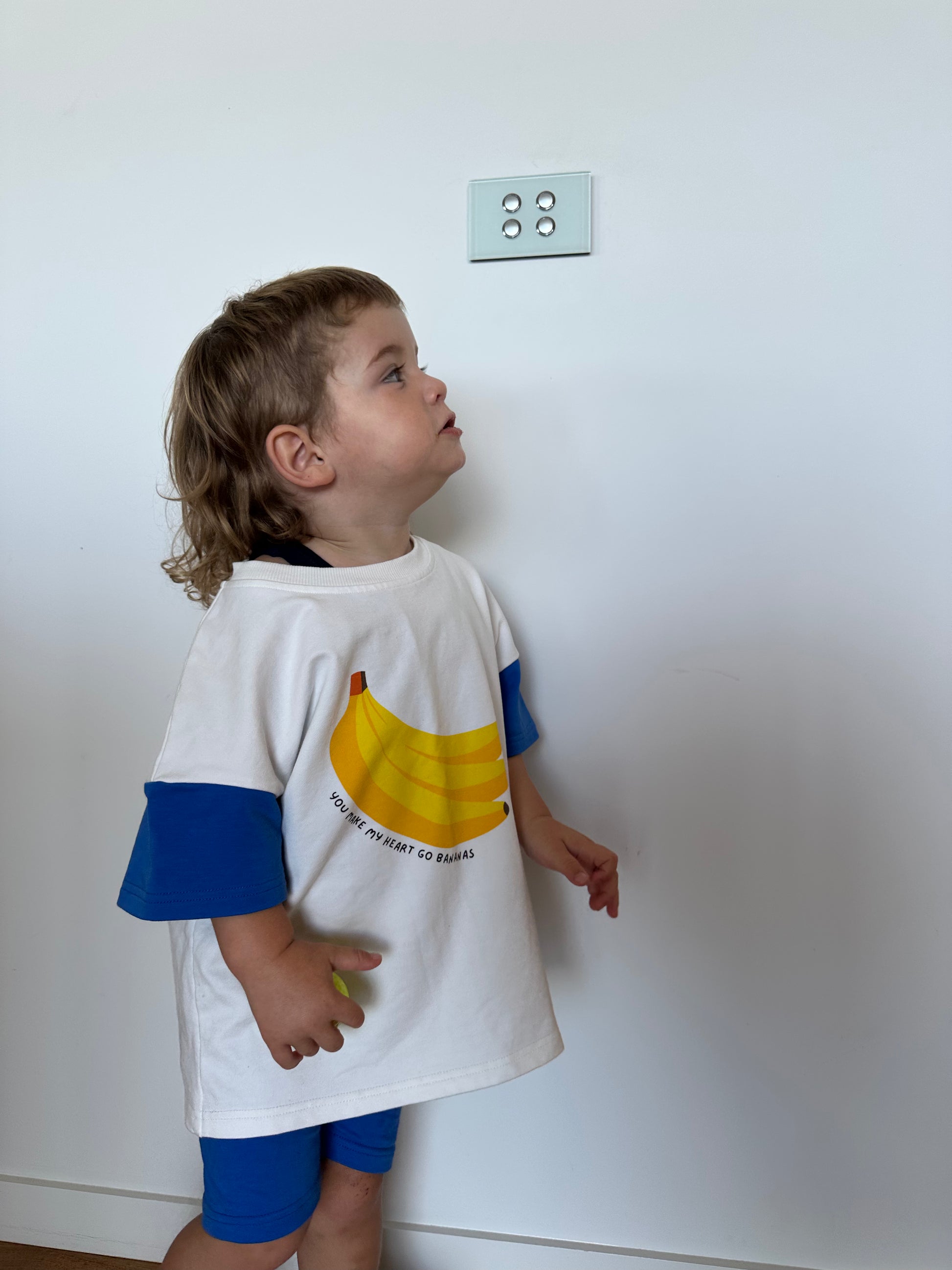 Child wearing a white t-shirt with a banana graphic and text, standing against a plain wall.