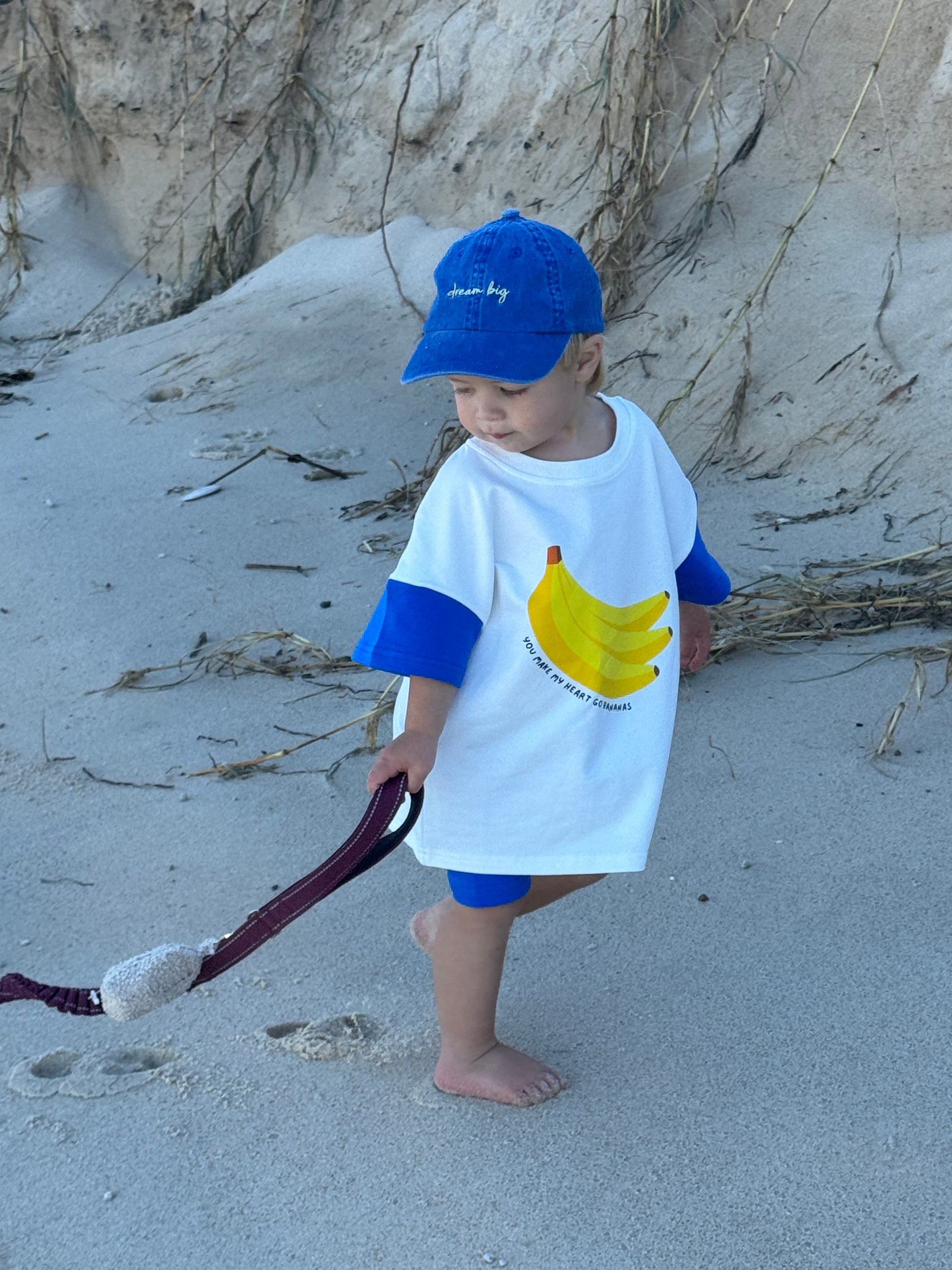 Child on a beach wearing a shirt with banana graphics and a blue cap.